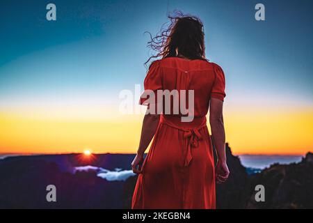 Beschreibung: Schöne braune Frau in rotem Kleid, die die sonnige Abendstimmung auf dem Pico do Ariero genießt. Verade do Pico Ruivo, Madeira Stockfoto