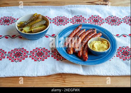 Eine schöne Aufnahme von gegrillten Würstchen, Kartoffelpüree, Gurken in Tongeschirr auf russische Stickerei Tischdecke gelegt Stockfoto