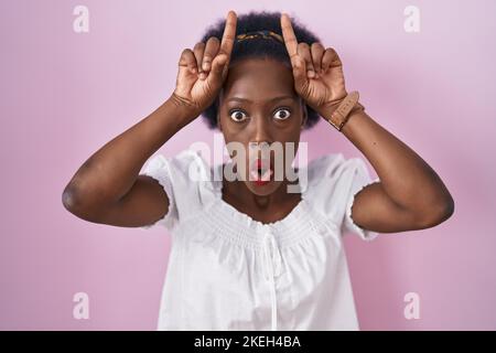 Afrikanische Frau mit lockigem Haar, das auf rosa Hintergrund steht und lustige Geste mit Finger über dem Kopf als Bullenhörner macht Stockfoto