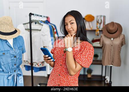 Junge hispanische Frau, die in einer Einzelhandelsboutique mit einem Smartphone arbeitet und selbstbewusst auf die Kamera blickt, lächelt mit gekreuzten Armen und erhobener Hand am Kinn. Th Stockfoto