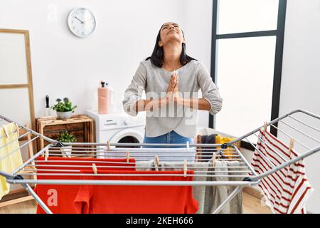 Junge hispanische Frau, die frische Wäsche auf die Wäscheleine legt, betteln und beten mit Händen zusammen mit Hoffnungsausdruck im Gesicht sehr emotional und verarrt Stockfoto