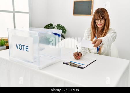 Junge hispanische Frau lächelt selbstbewusst und hält Stimmabgabe und schreibt in die Zwischenablage am Wahlkollegium Stockfoto