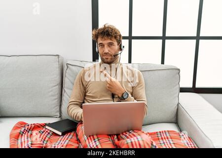 Ein junger, gutaussehender Mann mit Bart, der ein Headset trägt und von zu Hause aus mit dem Finger zur Seite zeigt und Werbung zeigt, ernst und ruhig Stockfoto