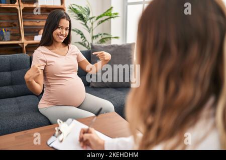 Junge Schwangere bei der Therapie-Sitzung mit einem Lächeln auf dem Gesicht, zeigt sich mit stolzen und glücklichen Fingern. Stockfoto