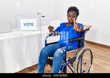 Junge afrikanerin, die auf einer Rollstuhlabstimmung sitzt, umhüllt die Wahlurne, zeigt den Finger nach oben und wütend, zeigt keine Geste Stockfoto