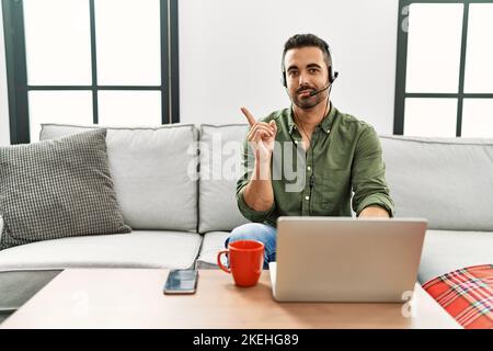 Junger hispanischer Mann mit Bart, der ein Callcenter-Agenten-Headset trägt und von zu Hause aus mit dem Finger zur Seite zeigt und Werbung zeigt, ernst Stockfoto