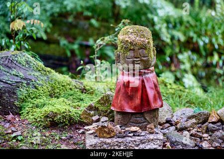 Moos-bedeckter Jizo mit rotem Kaminsims, der auf einem Fußweg in einem japanischen Kyoto-Tempelgarten in Japan mit geschlossenen Augen steht Stockfoto