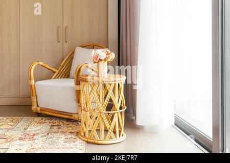 Rattanstuhl aus Holz mit grauem Kissen, farbigem Teppich, runder Tisch in der Ecke mit weißem Vorhang hinten an sonnigen Tagen Stockfoto