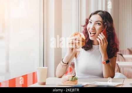 Eine Frau isst in einem Fast-Food-Café einen Hamburger und telefoniert mit einem Freund Stockfoto