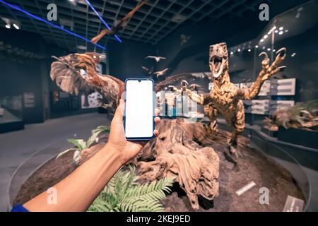 26. Juli 2022, Münster Natural History Museum, Deutschland: Besucherhand mit leerem Smartphone-Bildschirm bei der Ausstellung des furchterregenden gefiederten Dinosauriers de Stockfoto