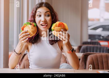 Glückliches Mädchen mit asiatischem und gemischtem Aussehen, das zwischen zwei Burgern oder einem Sandwich wählen kann. Ein Vegetarier - der andere mit Fleisch Stockfoto