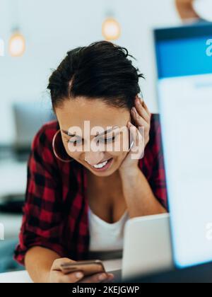 Eine Frau am Schreibtisch hält das Smartphone und starrt auf den Laptop, synchronisiert Daten zwischen Computer und Gadget im Büro mithilfe von Unternehmensgeräten Stockfoto