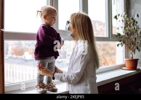 Seitenansicht eines kleinen Mädchens, das auf dem Fensterbrett in der Nähe des Kinderarztes steht und einen Kinderarzt in weißer Uniform ansieht. Stockfoto