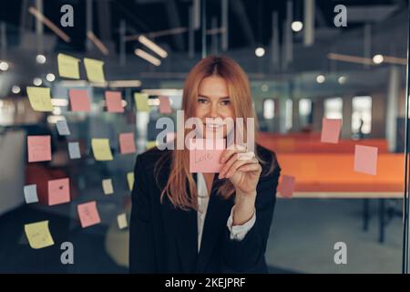 Lächelnde Geschäftsfrau zeigt Haftnotizen mit motivierenden Phrasen, während sie im Büro steht Stockfoto