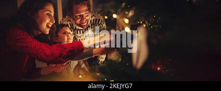 Dreiköpfige Familie bereitet Tannenbaum für Weihnachtsfeierlichkeiten zu Hause vor. Familie Dekoration Weihnachtsbaum zusammen zu Hause. Stockfoto