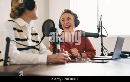 Weibliche Podcasterinnen lachen und haben eine tolle Zeit auf einer Audio-Übertragung in einem Home-Studio. Zwei glückliche Frauen, die einen Internet-Podcast für ihren SoC Aufnahmen Stockfoto