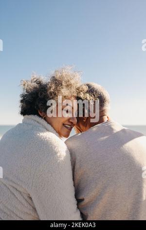 Rückansicht einer glücklichen älteren Frau, die lächelt, während sie mit ihrem Mann vor dem Meer sitzt. Rentnerpaar, das eine gute Zeit verbringt Stockfoto