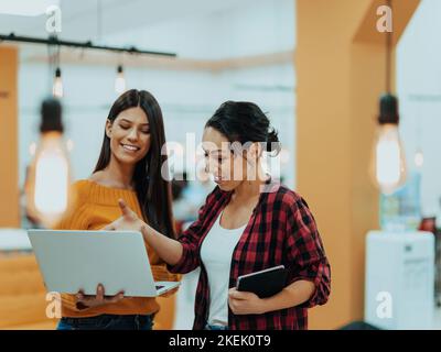 Porträt von zwei Geschäftsfrauen, die miteinander sprechen, während sie mit ihren Kollegen in einem modernen Geschäftsbüro mit Laptop und Tablet stehen Stockfoto