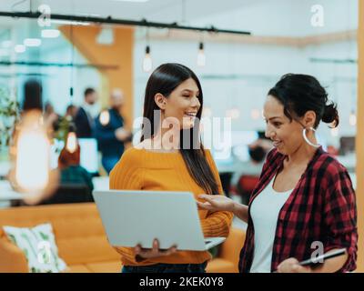 Porträt von zwei Geschäftsfrauen, die miteinander sprechen, während sie mit ihren Kollegen in einem modernen Geschäftsbüro mit Laptop und Tablet stehen Stockfoto
