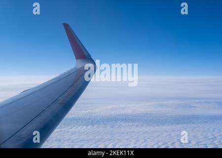 Der Flügel des Flugzeugs vor dem Hintergrund der weißen Wellenwolken und der Stratosphäre. Hintergrund des blauen Himmels mit flauschigen dichten Wolken. Himmelsverlauf. Stockfoto