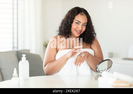 Zufriedene afroamerikanische mollige Dame im Handtuch, die Creme auf die Hand apponiert und nach der morgendlichen Dusche im Schlafzimmer sitzt Stockfoto