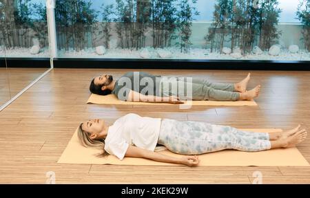 Fitness-Paar in Sportkleidung üben Yoga, während sie sich in Savasana oder Leichenhaltung im Yoga-Zentrum hinlegen. Shavasana ist eine wichtige Pose im restaurativen Yoga Stockfoto