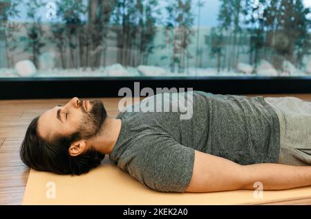 Savasana oder Leiche tot Pose. Der gutaussehende Mann entspannt sich in bewegungsloser Leichenhaltung mit geschlossenen Augen auf dem Boden des Meditationszentrums Stockfoto