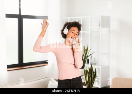 Eine fröhliche, tausendjährige afroamerikanische Frau in kabellosen Kopfhörern singt Lied, genießt Lieblingsmusik und tanzt Stockfoto