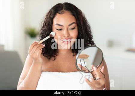 Junge schwarze Körper positive Dame hält Spiegel und Anwendung Rouge auf Gesicht mit Make-up-Bürste, tun täglich Make-up Stockfoto
