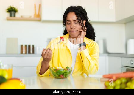 Gesunde Ernährung. Traurige afroamerikanische Frau, die frischen Gemüsesalat isst, am Esstisch in Kicthen sitzt, freien Platz Stockfoto