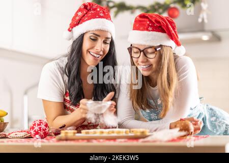 Eine glückliche Mutter und ihre fast Erwachsene Tochter machen zu Weihnachten Kuchen. Zwei fröhliche Frauen in Weihnachtsschürzen stauben Linn Kuchen mit Sug Stockfoto