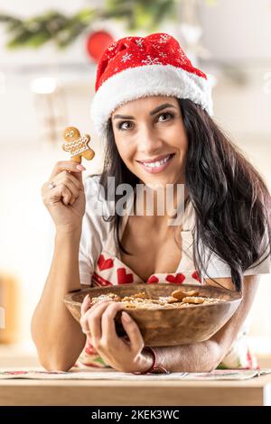 Eine junge Brünette in einer Weihnachtsschürze hat eine volle Schüssel Lebkuchen gebacken, sie hält einen davon zufrieden mit ihrer Arbeit in der Hand. Stockfoto