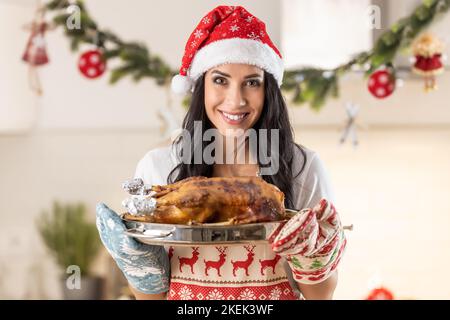 Zufriedene junge Frau in einer Weihnachtsschürze und Weihnachtsmütze mit einer Bratpfanne mit einer gebratenen Gans oder einem truthahn. Stockfoto