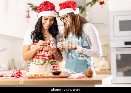 Eine Mutter und ihre Teenager-Tochter, die in Weihnachtsschürzen gekleidet sind, machen zu Hause in der Küche Weihnachtskekse. Stockfoto