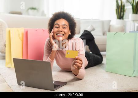 Lächelnde tausendjährige afroamerikanische Frau mit vielen Paketen zeigt, dass die Kreditkarte Laptop benutzt, auf dem Boden liegt Stockfoto