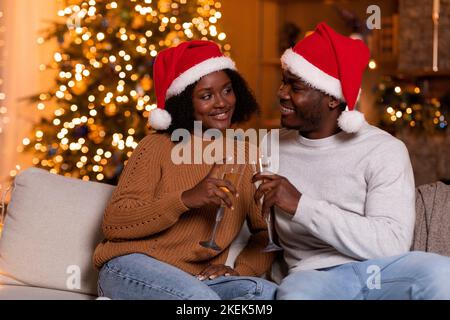 Lächelndes junges schwarzes Paar in Weihnachtsmützen jubelt mit einem Glas Champagner, genießt den Urlaub, sitzt auf dem Sofa Stockfoto
