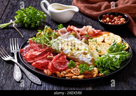 Herzhaft gebackener Brie mit Crackern, spanischem Salchichon, Salami-Würsten, dünn geschnittenem geräuchertem Schinken, grünen Oliven, Salat, Erdnüssen, Pecan auf schwarzer Platte Stockfoto