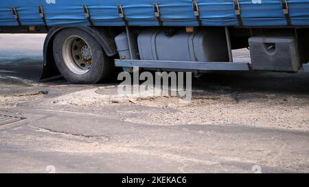 Nahaufnahme, sehr schlechte Straßenabdeckung, viele Gruben, gekrümmter Asphalt, ein großer LKW passiert langsam ein Stück Straße in Verfall. Die Straße muss repariert werden. Hochwertige Fotos Stockfoto