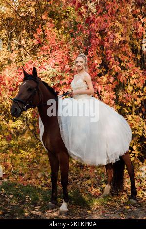 Schöne Braut in weißem Kleid sitzt auf einem Pferd im Herbstwald Stockfoto