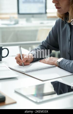 Nahaufnahme einer jungen Frau, die Pläne in ihr Notebook geschrieben hat, während sie am Arbeitsplatz im Büro sitzt Stockfoto