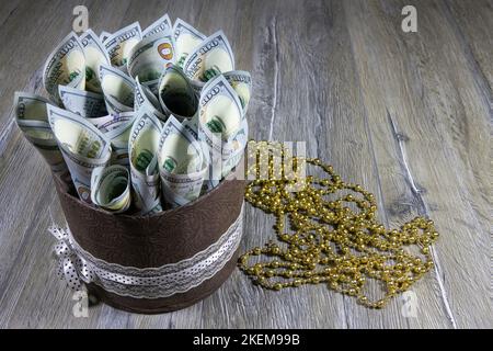 Geschenkkarton in Form eines Zylinders mit einhundert Dollarscheinen, die auf einem grauen Holztisch in ein Rohr gerollt werden. Banken und Finanzwesen Stockfoto