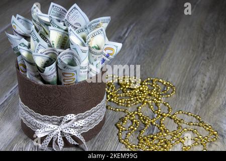 Geschenkkarton in Form eines Zylinders mit einhundert Dollarscheinen, die auf einem grauen Holztisch in ein Rohr gerollt werden. Banken und Finanzwesen Stockfoto