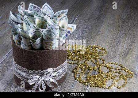 Geschenkkarton in Form eines Zylinders mit einhundert Dollarscheinen, die auf einem grauen Holztisch in ein Rohr gerollt werden. Banken und Finanzwesen Stockfoto