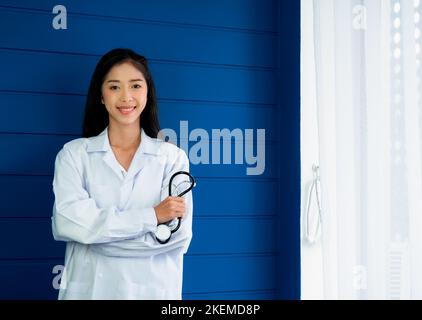 Lächelnd hübsche asiatische Frau Arzt Porträt steht auf blauem Holz Hintergrund in der Nähe des Vorhangs Fenster in der medizinischen Praxis. Selbstbewusst asiatische junge weibliche tun Stockfoto