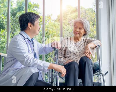 Lächelnd positiver asiatischer Mann Arzt im weißen Mantel spricht mit Sorgfalt zu älteren älteren älteren weiblichen Patienten, die in einem Rollstuhl in der medizinischen Praxis in der Zusammenarbeit sitzen Stockfoto