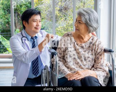 Lächelnd positiver asiatischer Mann Arzt im weißen Mantel spricht mit Sorgfalt zu älteren älteren älteren weiblichen Patienten, die in einem Rollstuhl in der medizinischen Praxis in der Zusammenarbeit sitzen Stockfoto