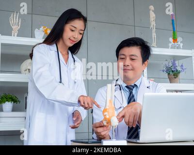 Zwei asiatische Ärzte in weißen Uniformen unterhalten sich und arbeiten im medizinischen Büro zusammen. Der Erwachsene männliche Arzt berät junge weibliche Arzt über die Stockfoto