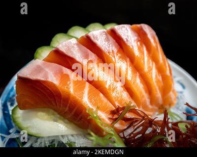 Sashimi aus frischem Lachs, berühmtes japanisches Essen. Fünf orangefarbene rohe Fischstücke, serviert mit Gurken auf Keramikplatte auf dunklem Hintergrund. Nahaufnahme SA Stockfoto