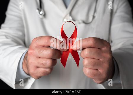 Nahaufnahme eines Arztes, der ein rotes Band mit beiden Händen hält, Symbol des Welt-AIDS-Tages. Unterstützung von Gesundheitspersonal und HIV-infizierten Menschen. Prävention und Stockfoto