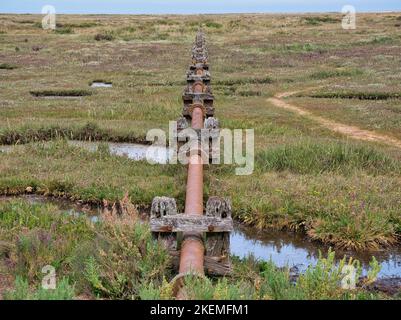 Eine rostende Drainage auf verwitterten Holzstützen bei Stiffkey Salt Marshes auf dem North Norfolk Coast Path. Das Rohr verläuft über die Sümpfe. Stockfoto
